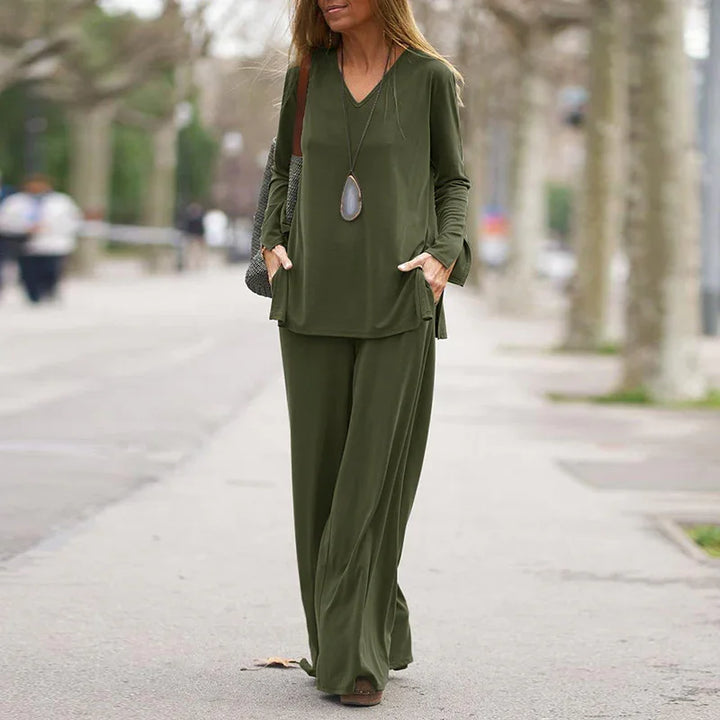 Woman wearing olive green loose fit pants and long sleeve top with pendant necklace walking on tree-lined sidewalk