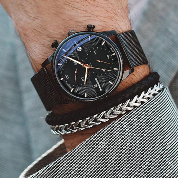 Black Bimond wristwatch with mesh strap and silver chain bracelet on a wrist in checkered shirt