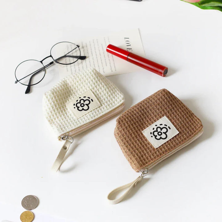 Two small waffle-textured zipper pouches in cream and brown with flower logos on a white table beside glasses, lipstick, and coins