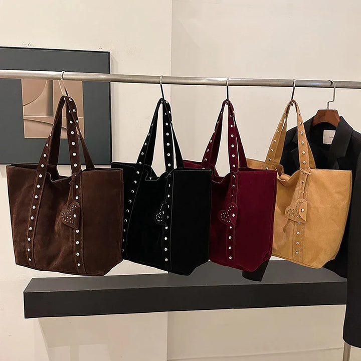 Four suede tote bags in brown, black, burgundy, and tan with heart-shaped studs hanging on a metal rack