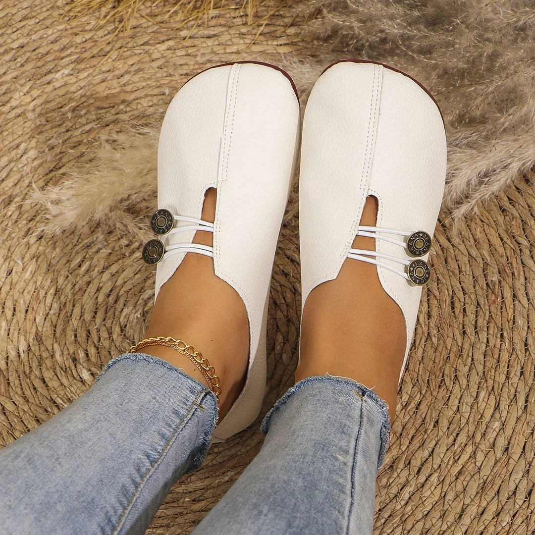 White leather slip-on shoes with button accents worn with light blue jeans and gold anklet on woven rug