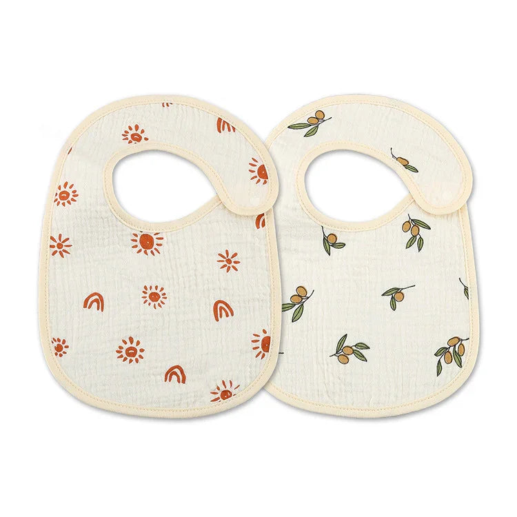Two cream-colored baby bibs with snap closures, one with orange sun and rainbow prints, the other with green and yellow berry prints