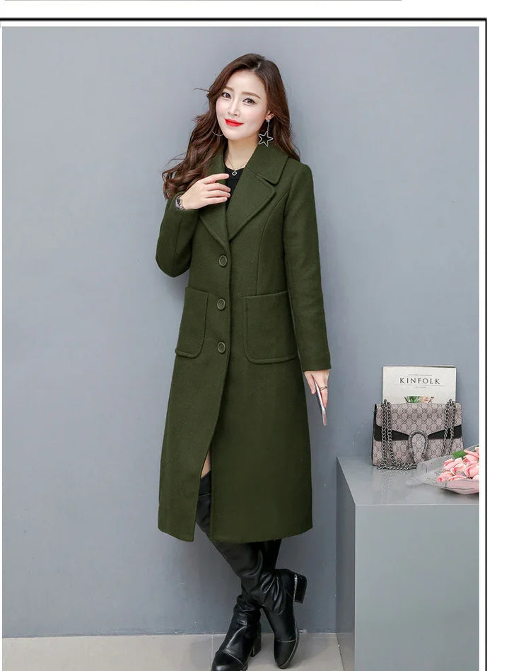 Woman modeling long dark green wool coat with large pockets and black boots against gray wall