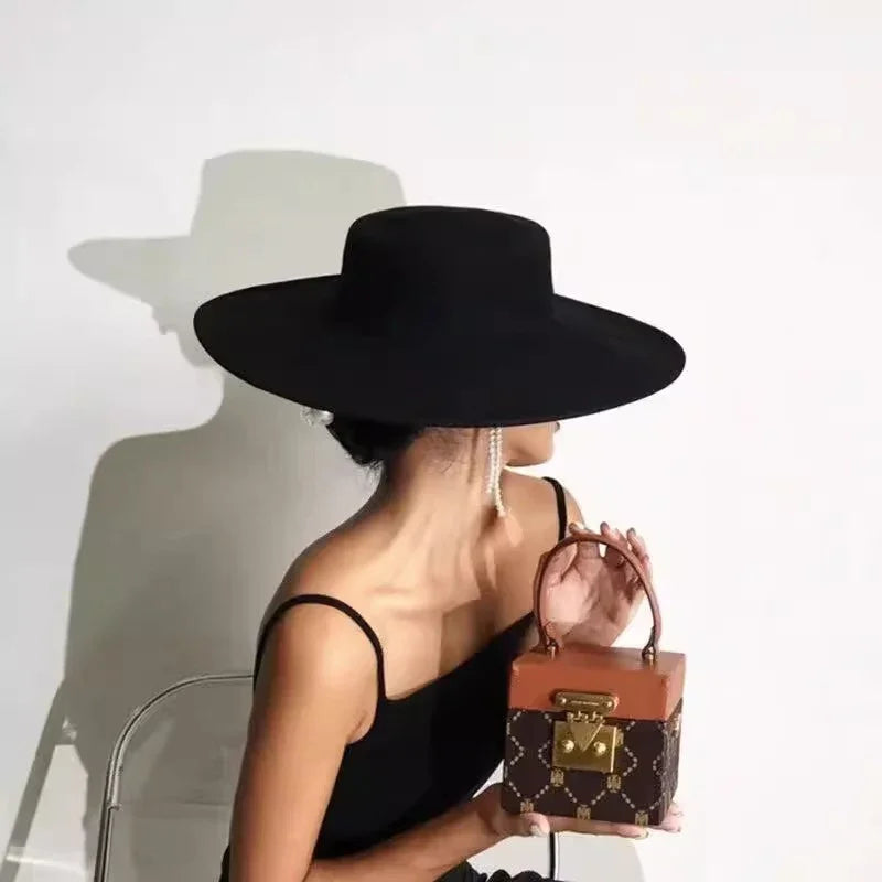 Woman in black dress and wide-brimmed black hat holding a brown monogram handbag against white background