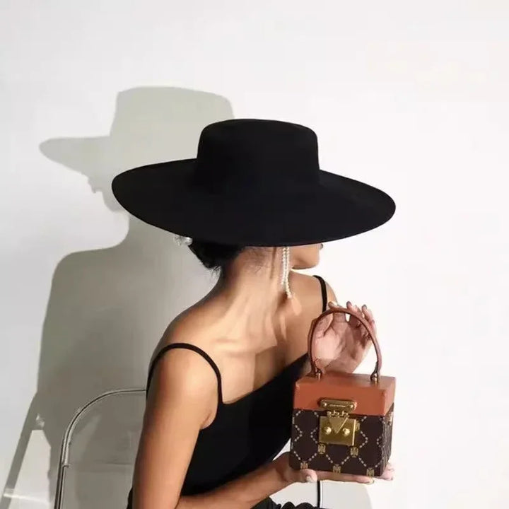 Woman in black dress and wide-brimmed black hat holding a brown monogram handbag against white background