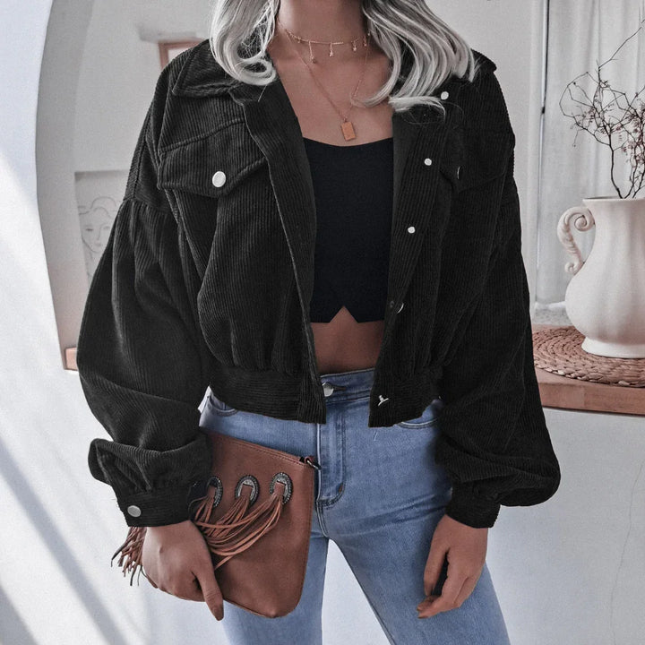 Woman wearing black cropped corduroy jacket and blue jeans holding brown fringe clutch bag indoors