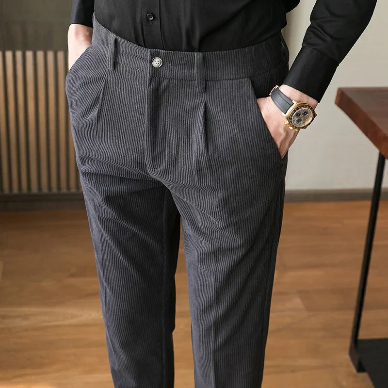Man wearing dark grey corduroy trousers with pleats and black shirt, hands in pockets, gold watch