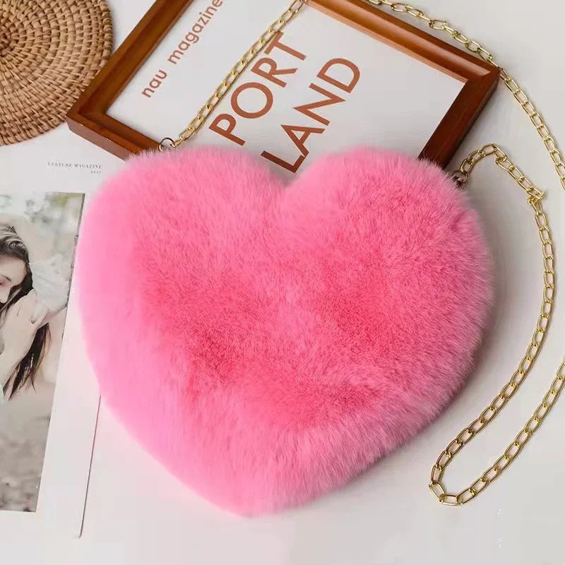Pink fluffy heart-shaped purse with gold chain strap on white surface with magazine and photo