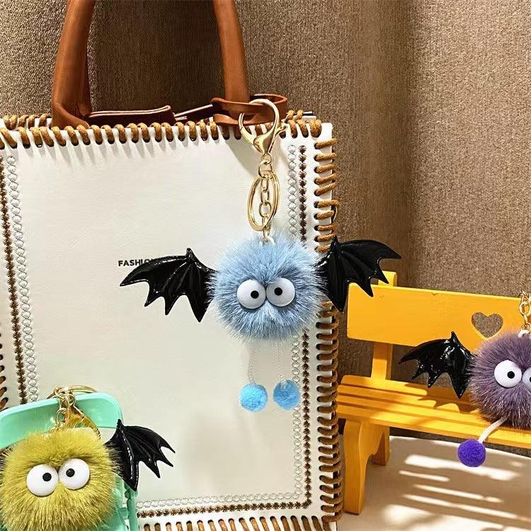 Fluffy bat keychains with big eyes and black wings hanging on white bag and yellow bench