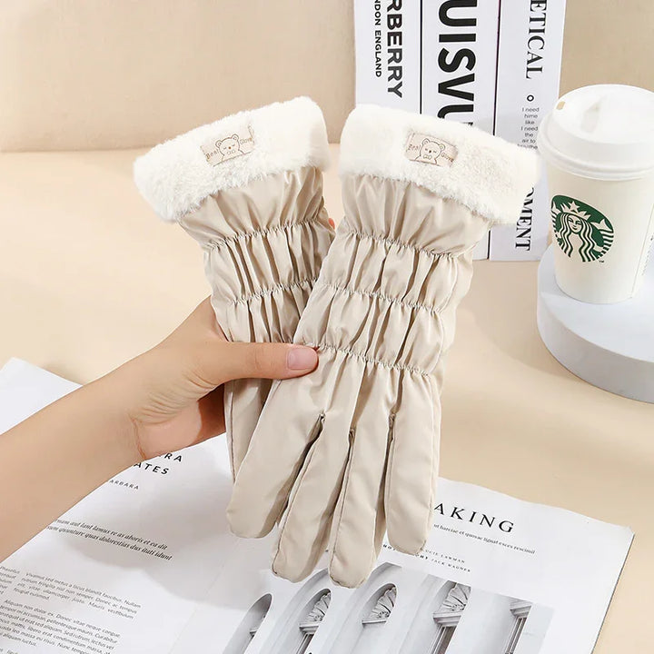 Beige winter gloves with white faux fur cuffs featuring a bear patch, held over a magazine and beside a Starbucks cup