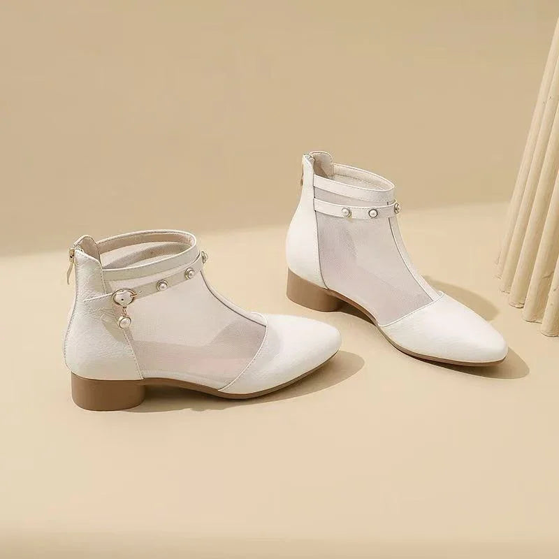 White ankle boots with pointed toes, mesh panels, decorative pearl buttons, and low brown heels on beige background