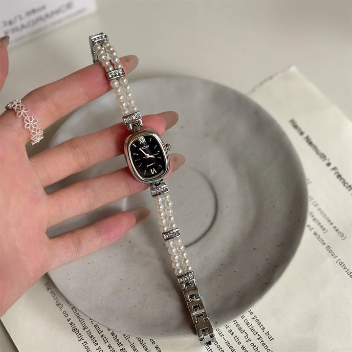 Elegant women's pearl bracelet watch with black dial and rhinestone accents, held over gray ceramic plate