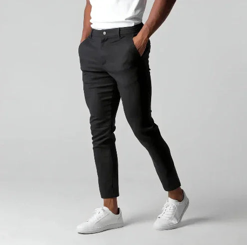 Man wearing slim fit black trousers and white sneakers with hands in pockets on grey background