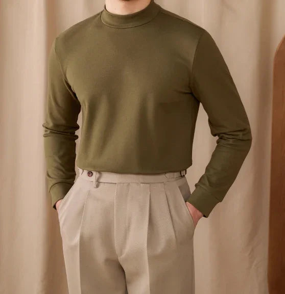 Man wearing olive green long sleeve mock neck shirt and beige pleated trousers with hands in pockets