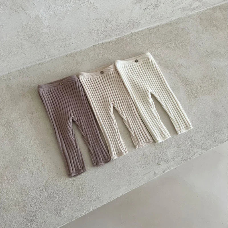 Three pairs of ribbed knit baby leggings in mauve, beige, and white laid out on a textured floor