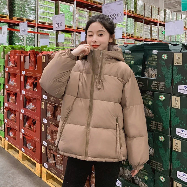 Young woman in a beige puffer jacket winking, standing in a grocery warehouse aisle with snacks on shelves