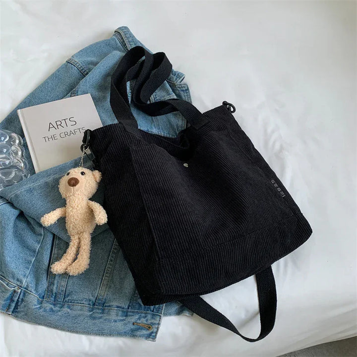 black corduroy tote bag with plush bear keychain on blue denim jacket and book on white surface