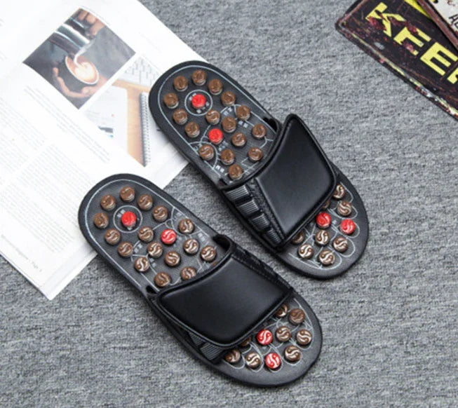 black foot massage slippers with red and brown acupressure nodes on gray carpet near open magazine