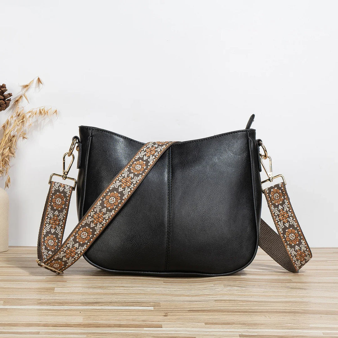 Black leather crossbody bag with adjustable floral-patterned fabric strap on wooden surface