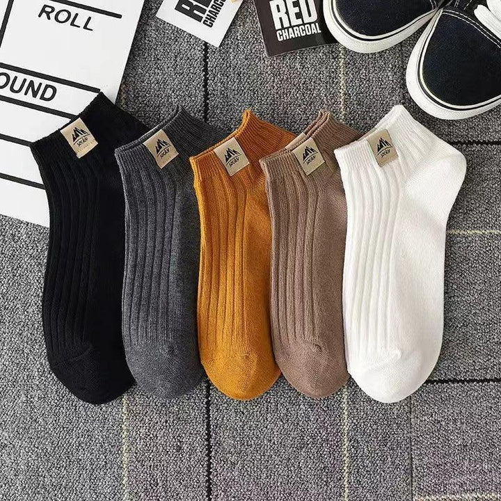 Set of five ribbed ankle socks in black, gray, mustard, brown, and white on carpet background with casual sneakers