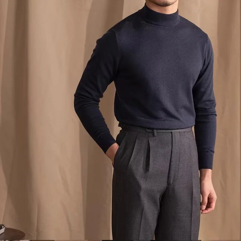 Man wearing navy blue turtleneck sweater and gray pleated trousers against beige background