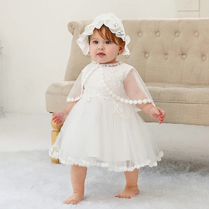 Baby girl wearing a white lace dress and bonnet standing barefoot on a fluffy rug in front of a beige tufted sofa