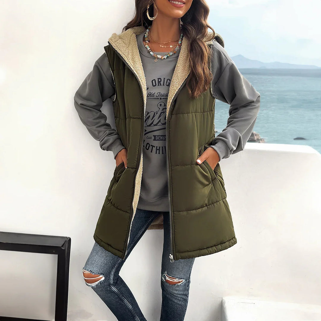 Woman wearing olive green hooded puffer vest with sherpa lining, gray sweatshirt, and ripped jeans by sea