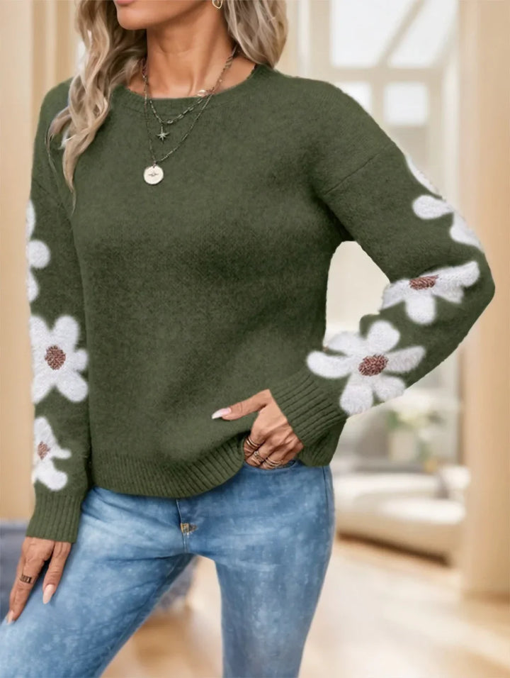 Woman wearing olive green sweater with white daisy floral sleeves and blue jeans in cozy indoor setting