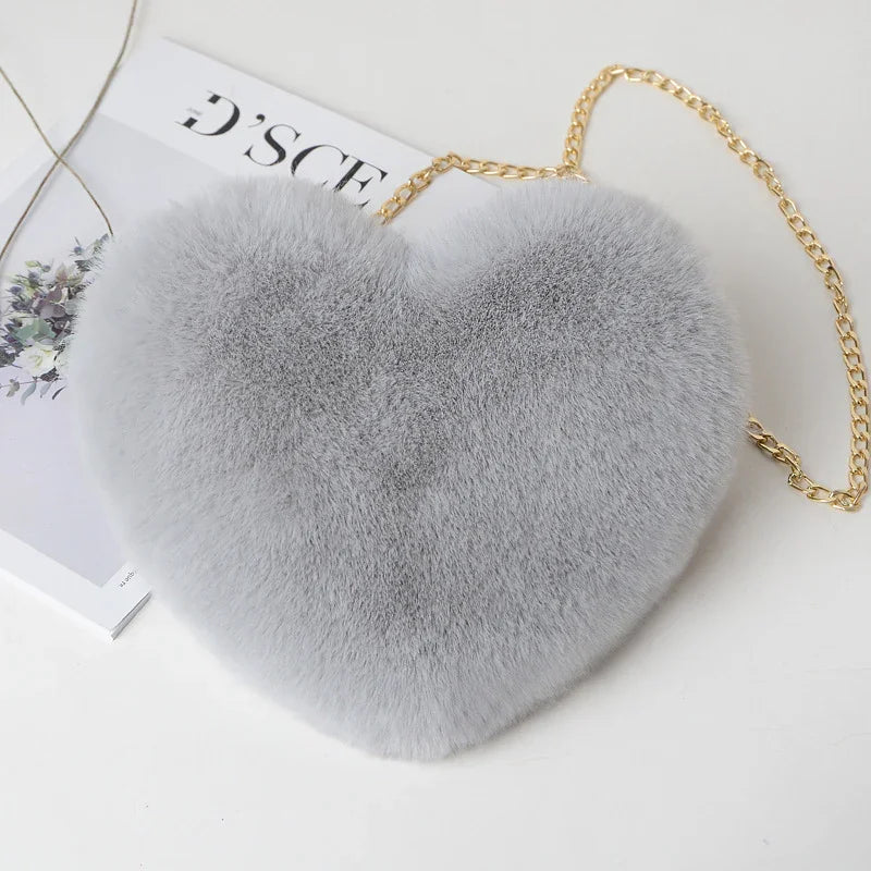 Gray heart-shaped faux fur handbag with gold chain strap on white surface with magazine