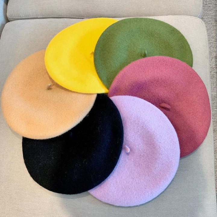 Six colorful wool berets in yellow, green, pink, light purple, black, and beige arranged on a beige fabric surface