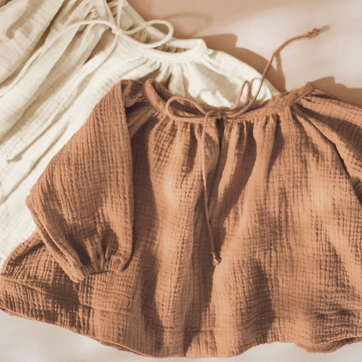 Two textured long-sleeve baby blouses in beige and brown with tie necks on a soft background