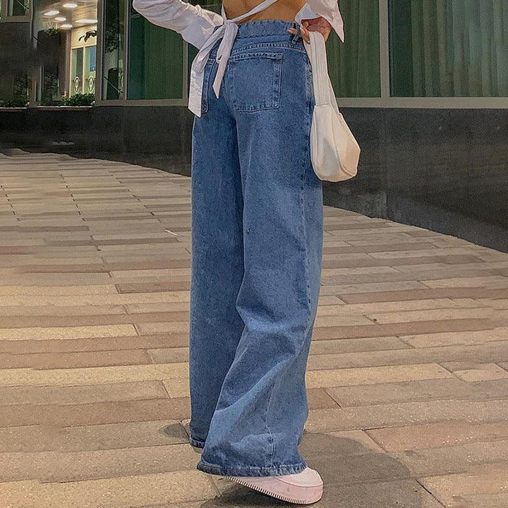 Woman wearing loose wide-leg blue jeans and white sneakers, standing on urban tiled pavement with a small white handbag