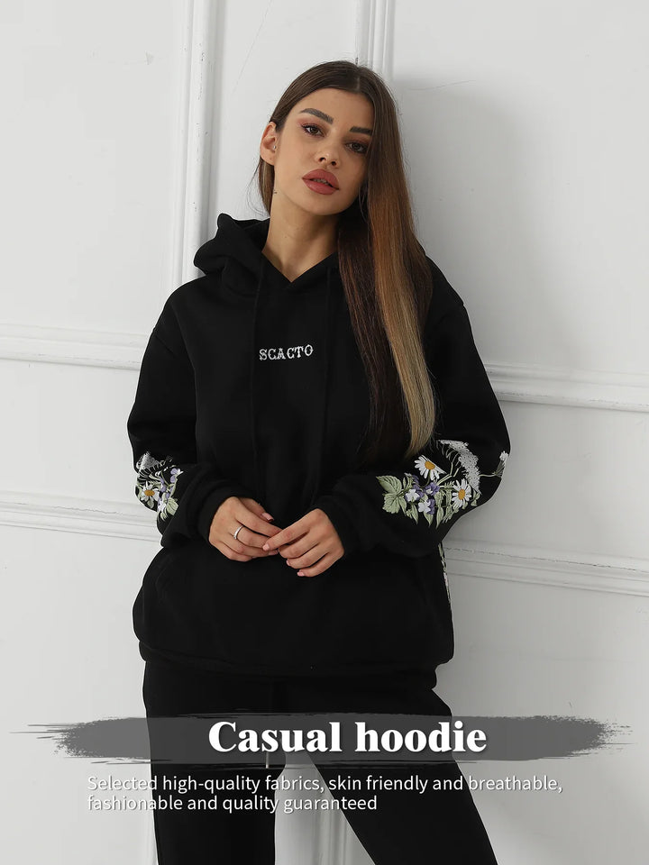 Woman wearing black casual hoodie with floral sleeves and SCAGTO logo, standing against white wall
