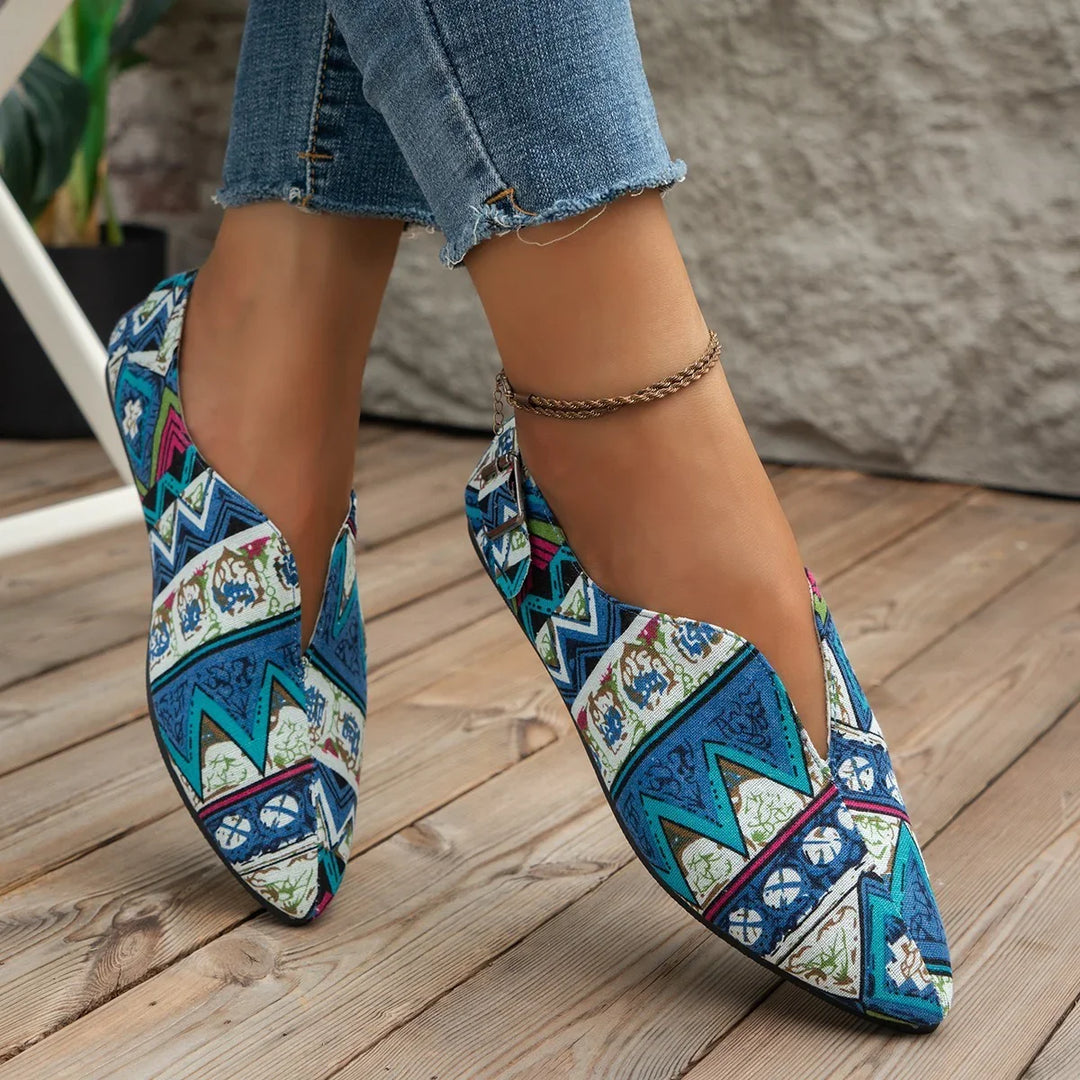 Close-up of feet wearing colorful blue patterned slip-on shoes with pointed toes and ankle bracelet on wooden floor