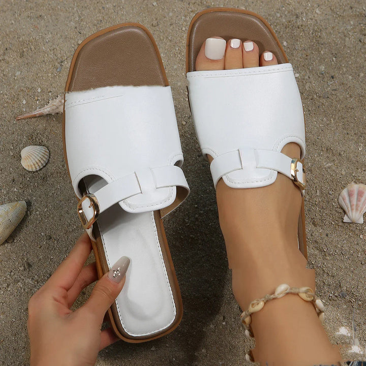 White leather slide sandals on sandy beach with seashells, featuring gold buckle and shell anklet