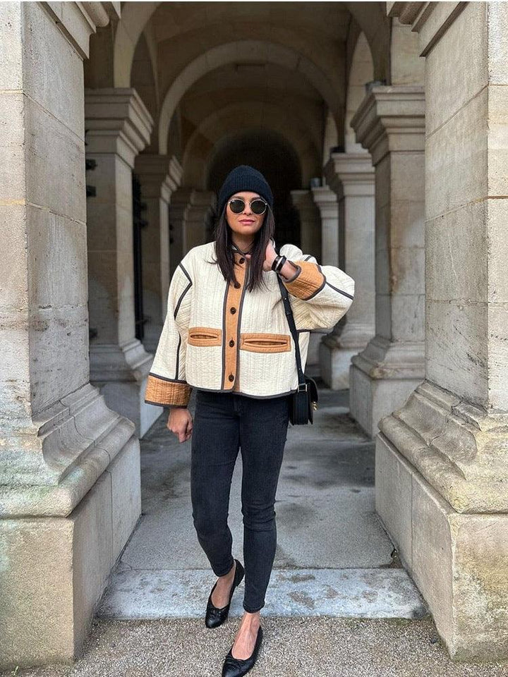 Woman wearing beige and brown jacket, black beanie, sunglasses, black skinny jeans, and flats in stone archway