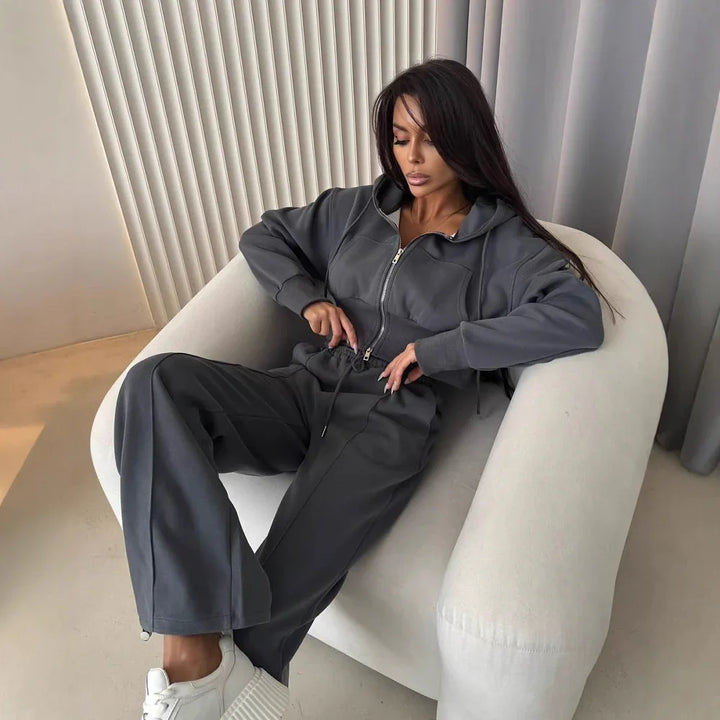 Woman sitting in white modern chair wearing grey zip-up hoodie, matching sweatpants, and white sneakers
