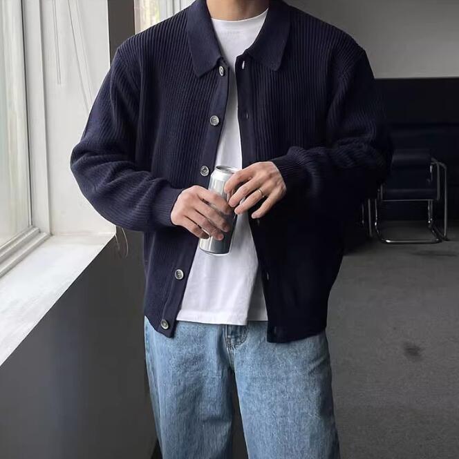 Man wearing black ribbed jacket over white t-shirt and blue jeans holding a beverage can indoors near window