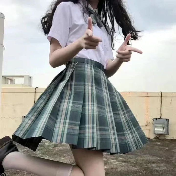 Person wearing a white blouse and green plaid pleated skirt posing with finger guns outdoors
