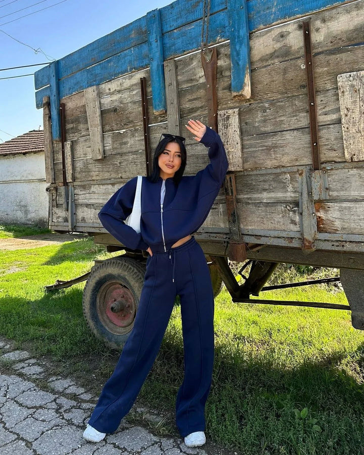 Woman in navy blue tracksuit and white sneakers posing by rustic wooden trailer outdoors