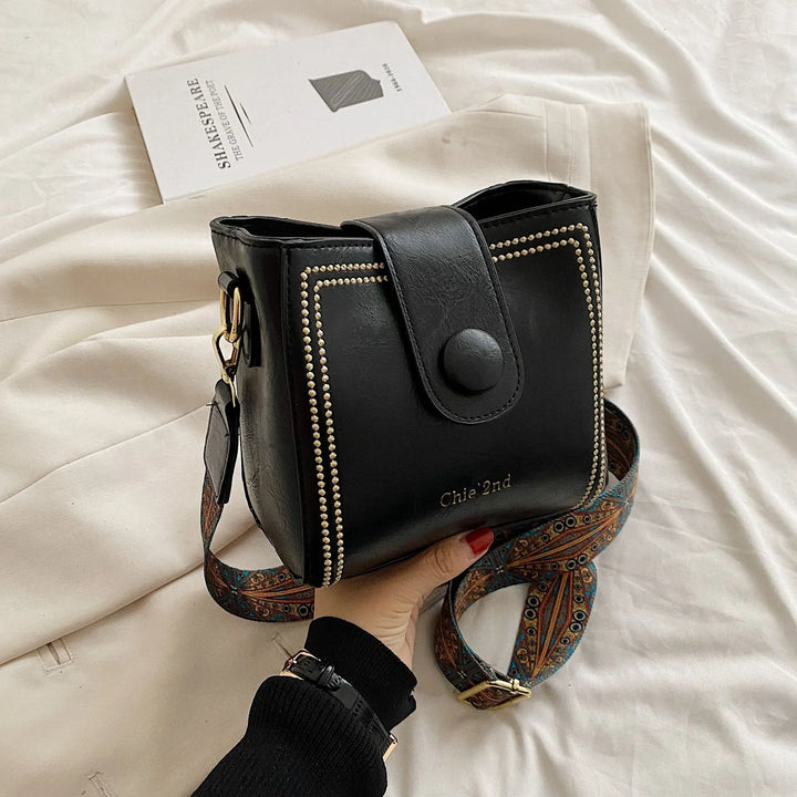 Hand holding black leather Chie 2nd crossbody bag with decorative stitching and colorful strap on white fabric