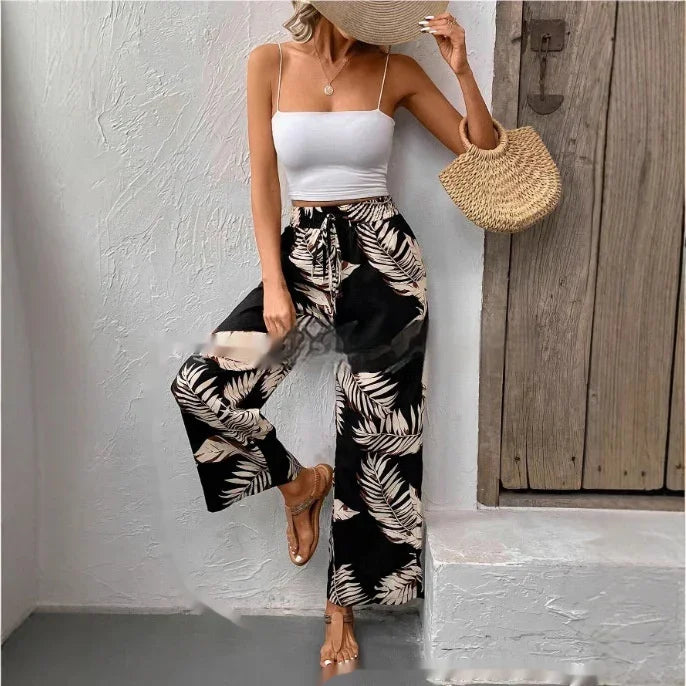 Woman wearing white spaghetti strap crop top, black wide-leg pants with beige leaf print, holding straw hat and woven bag