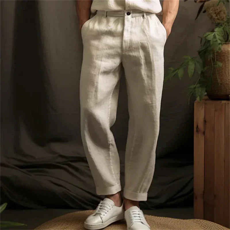 Man wearing beige linen pants and white sneakers standing indoors with plants and wooden furniture