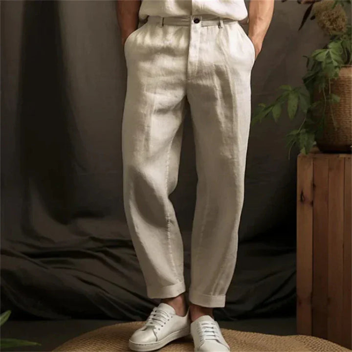 Man wearing beige linen pants and white sneakers standing indoors with plants and wooden furniture