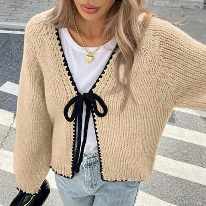 Woman wearing beige knitted cardigan with black trim and bow, white t-shirt, blue jeans, and gold heart necklace