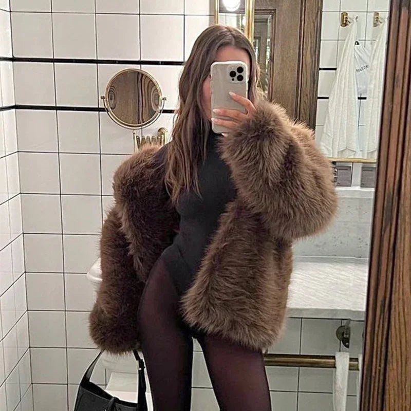 Woman in brown faux fur coat and black bodysuit taking mirror selfie in tiled bathroom