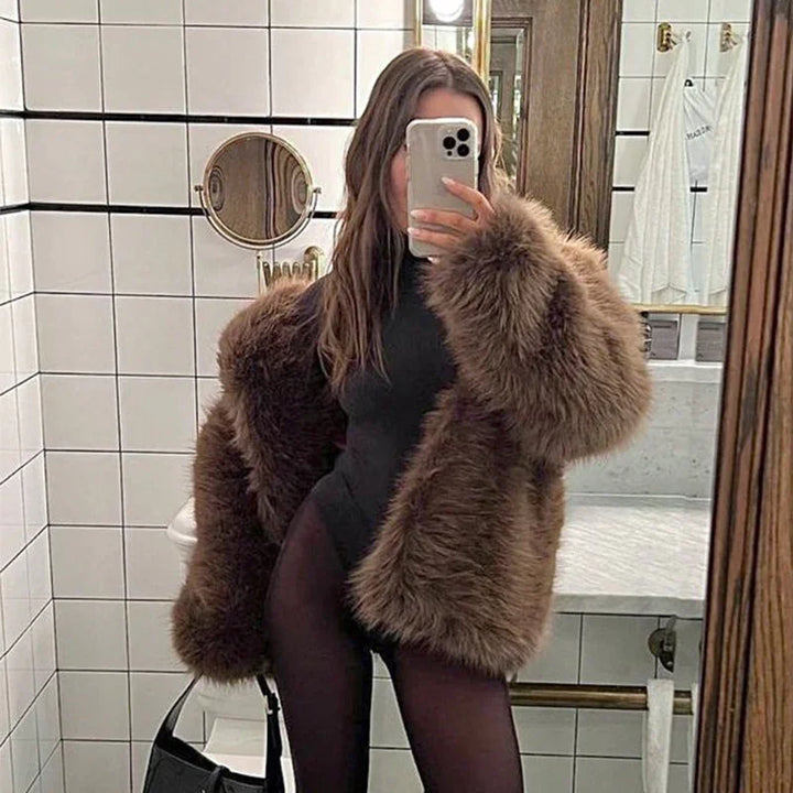 Woman in brown faux fur coat and black bodysuit taking mirror selfie in tiled bathroom
