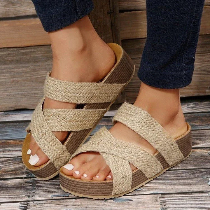 Beige woven platform sandals with crisscross straps worn with dark pants on wooden floor