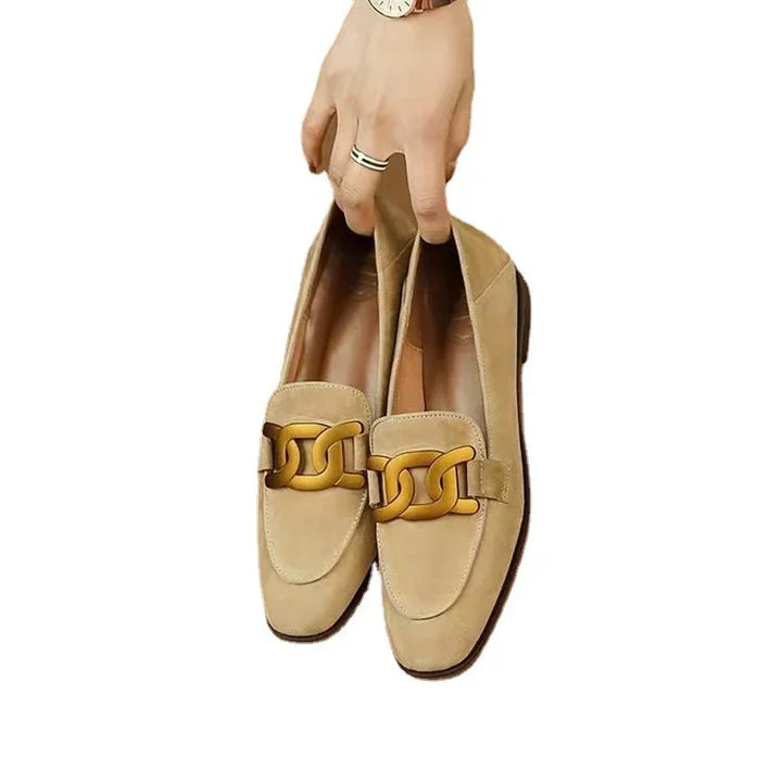 Hand holding beige suede loafers with large gold chain detail on white background