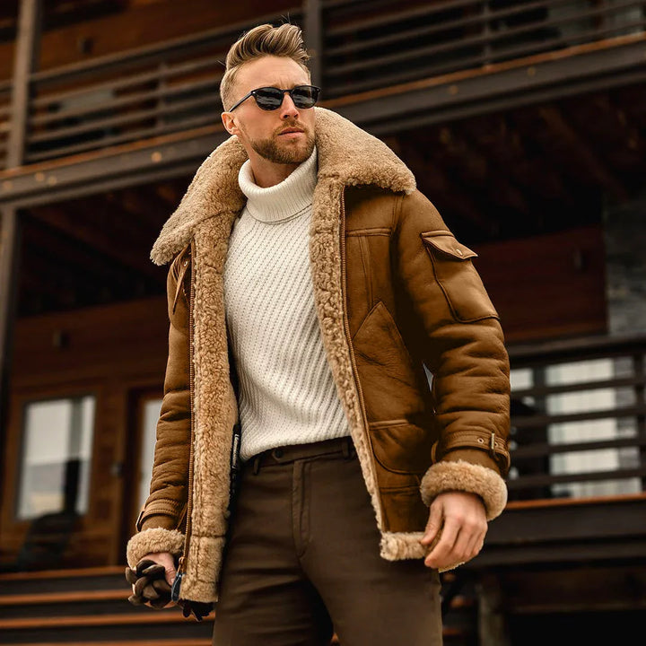 Man wearing brown shearling jacket, white turtleneck sweater, and sunglasses in front of wooden cabin